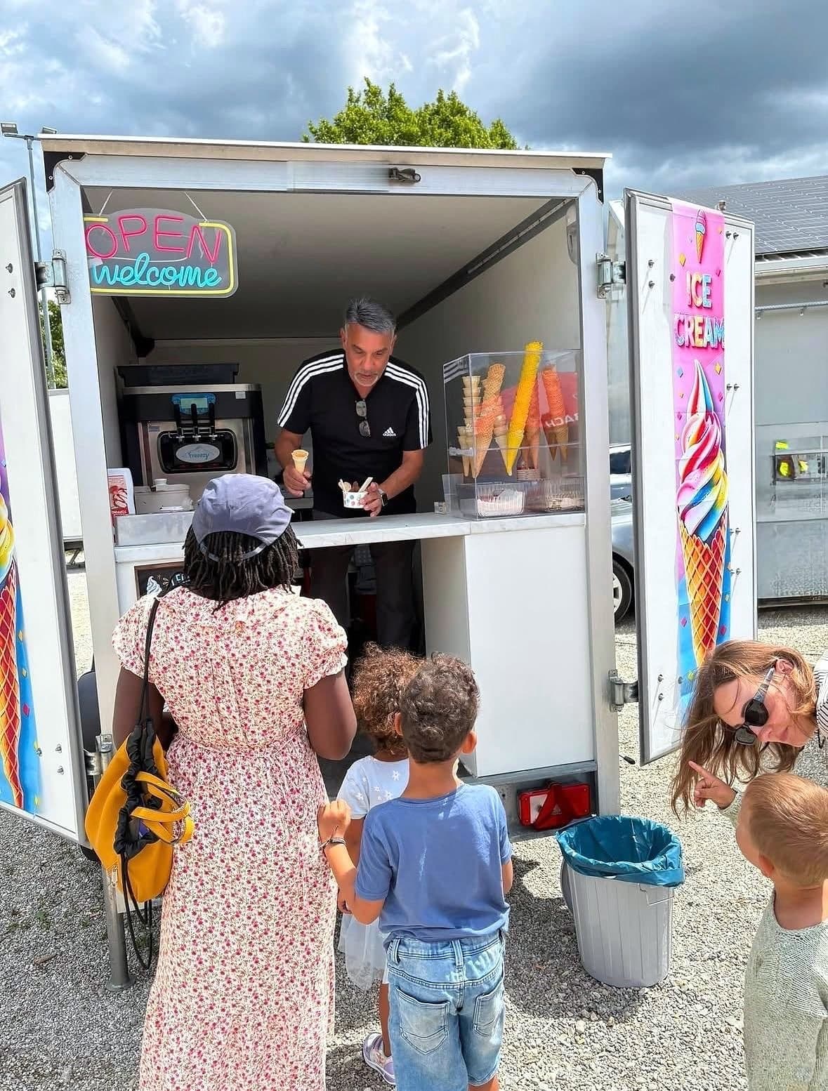 Street Food Festival Wasserburg am Inn: Mondy reicht an der Theke seines geöffneten Softeis-Wagens ein Eis heraus, darüber leuchtet das OPEN-welcome-Schild, davor wartet eine Familie