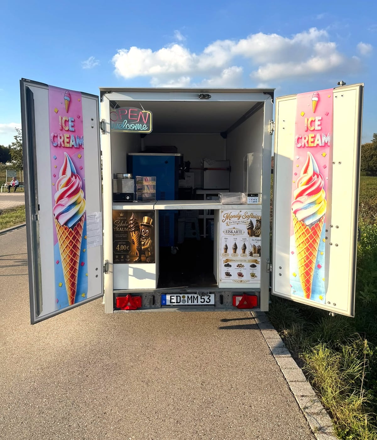 Der weiße Softeis-Anhänger von Mondy Softeis mit weit geöffneten Türen: an den Türinnenseiten die ICE-CREAM-Plakate, über der Theke das OPEN-welcome-Neonschild, rechts die Eiskarte von Mondy Softeis